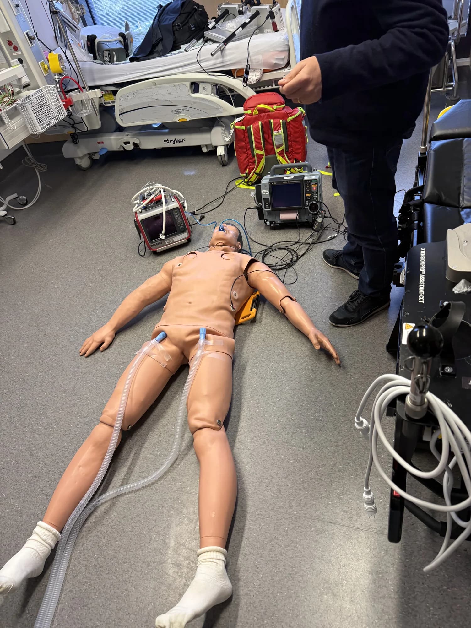 An ECPR training mannequin with cannulation tubing, monitoring equipment, and a transport stretcher nearby