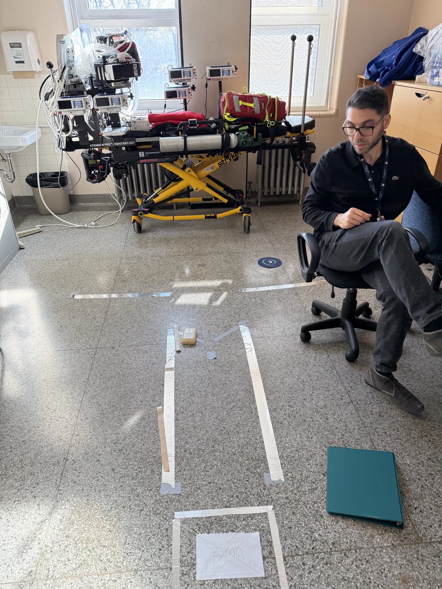 A colleague beside the equipped transport stretcher, with a taped equipment layout on the floor